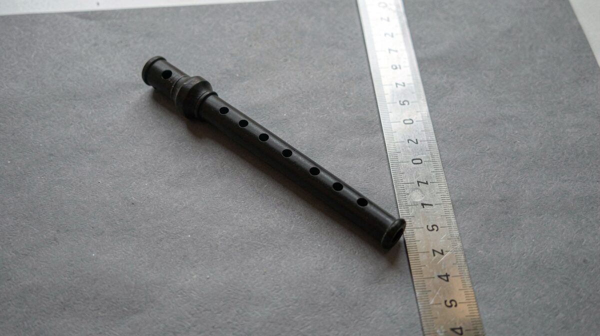A blackwood bagpipe chanter lying on a matte gray paper sweep beside a steel ruler, lit by two diffused lights at forty-five degrees, photographed square from above.